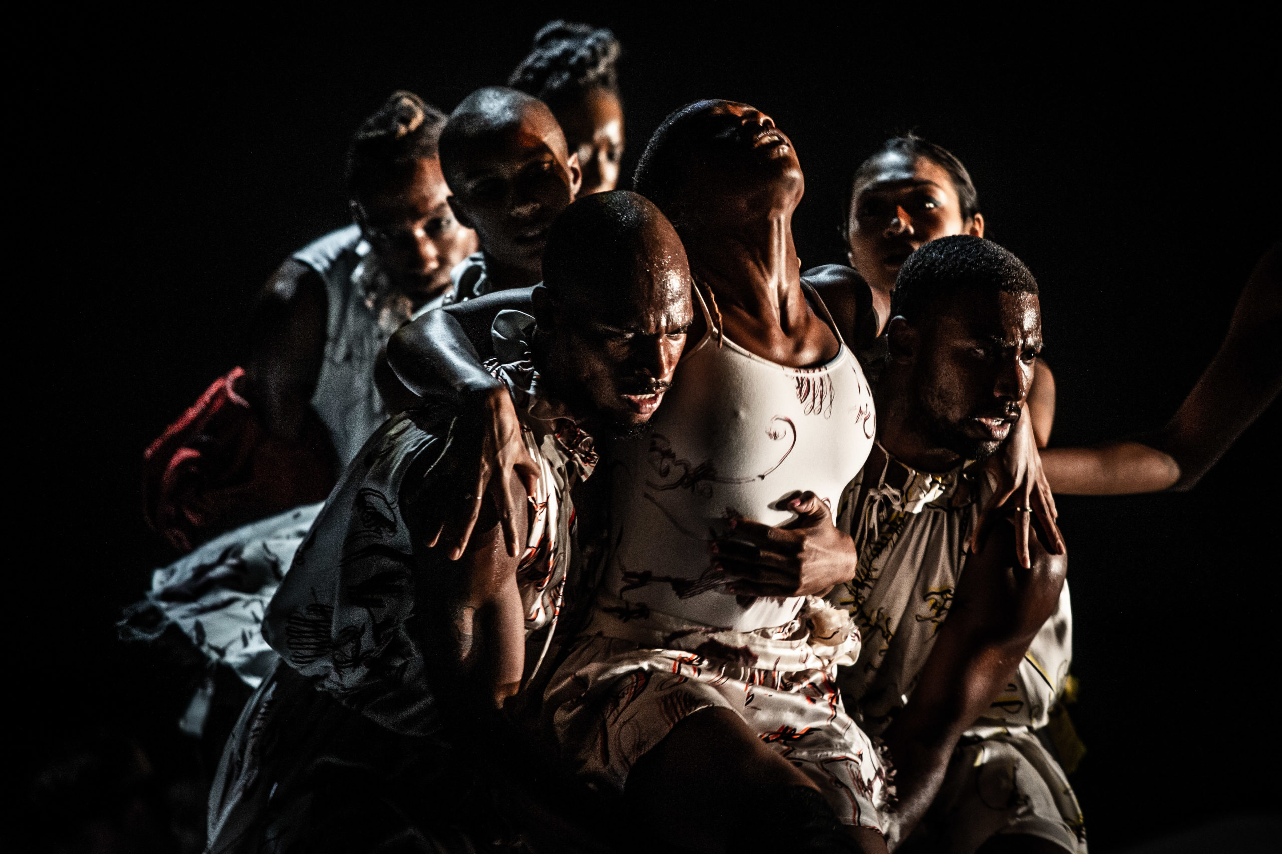 A dancer tips her head back, in agony or exhaustion or ecstasy, as the dancers on either side of her carry her forward, tucked under her shoulders with arms around her waist. Four more dancers are blurry but follow close behind, their limbs blocked by the procession before them.