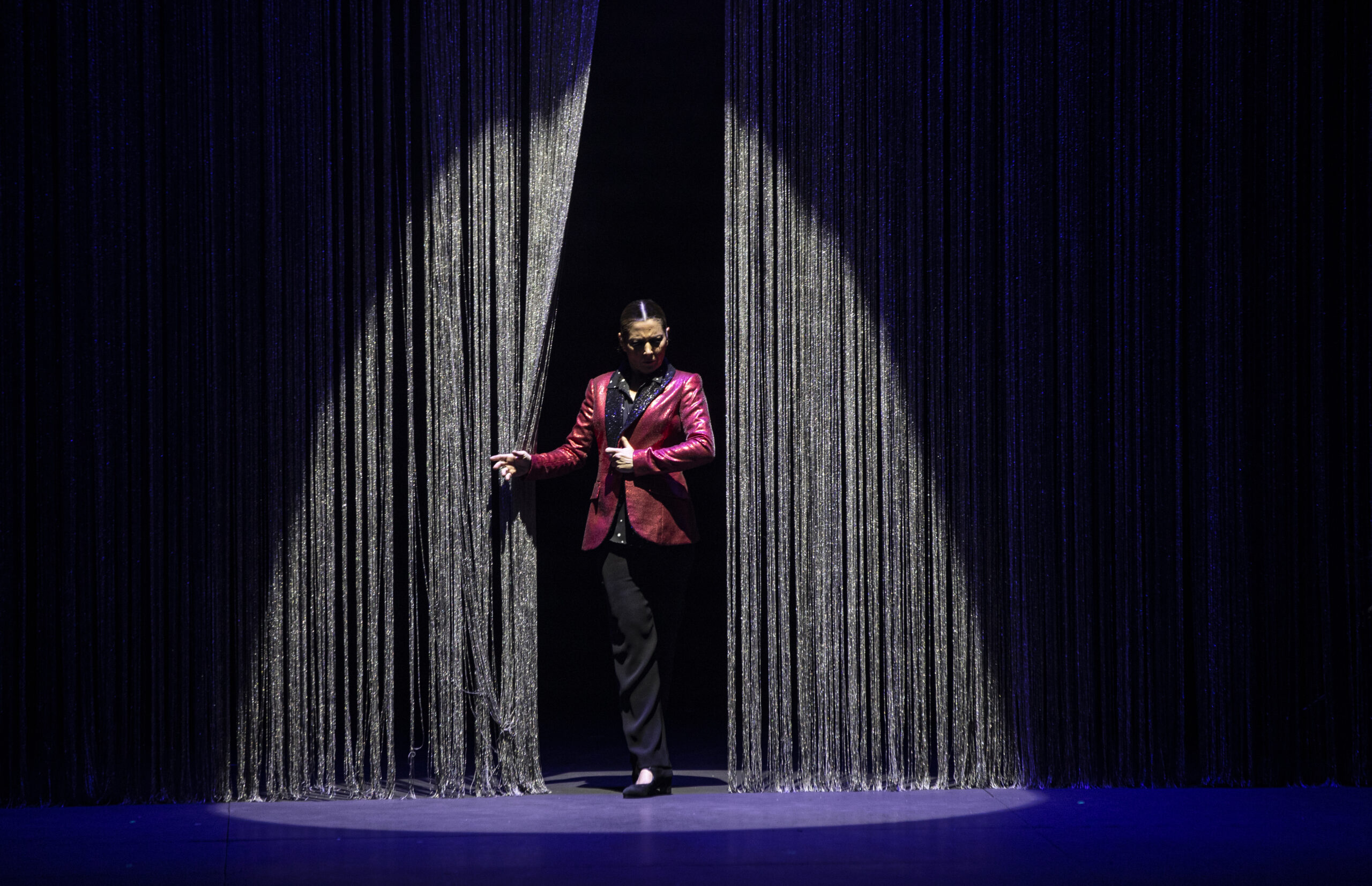 Sara Baras emerges into a spotlight from between a silver curtain. She wears a red suit jacket over a sparkling black shirt and dark trousers. Her chin dips toward her chest, one hand alighting at her sternum while the other brushes the silvery curtain aside.