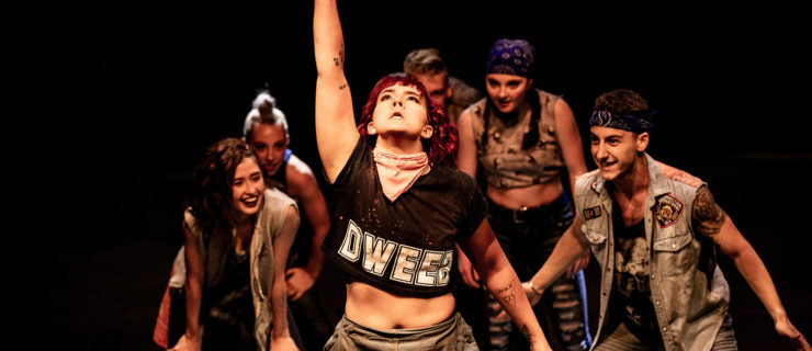 Dancers in street clothes gather behind a female dancer as she points to the sky with one finger.