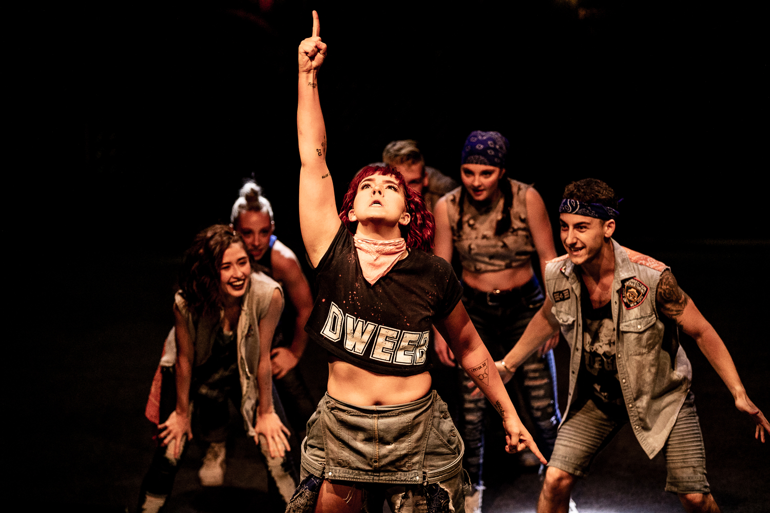 Dancers in street clothes gather behind a female dancer as she points to the sky with one finger.