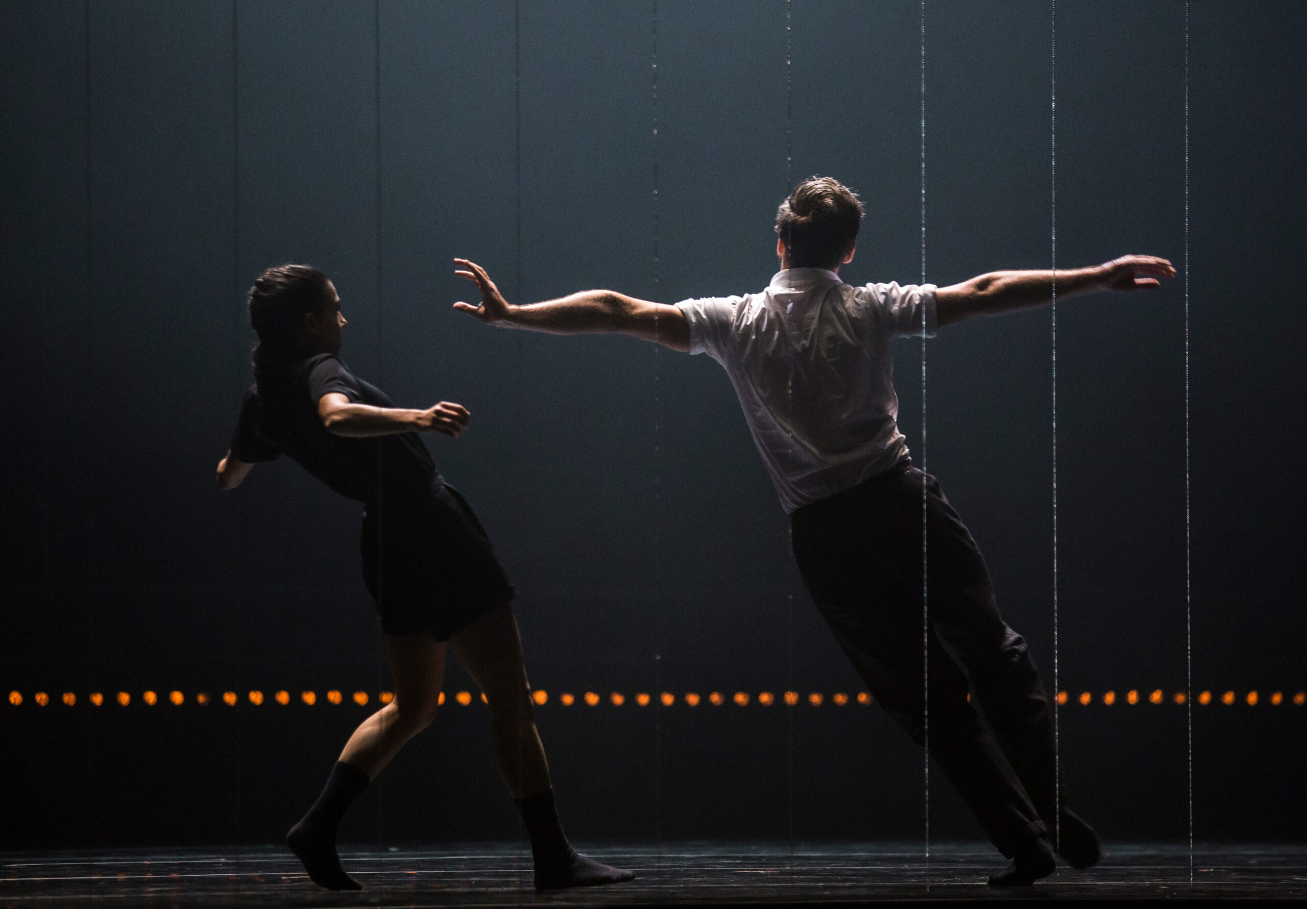 Two dancers face upstage, leaning off balance to their left. One outstretches their arms straight side, pulling toward their left hip. To their left, a dancer facing them rocks onto their back foot, shoulders up as they avoid the other dancer's extended hand.