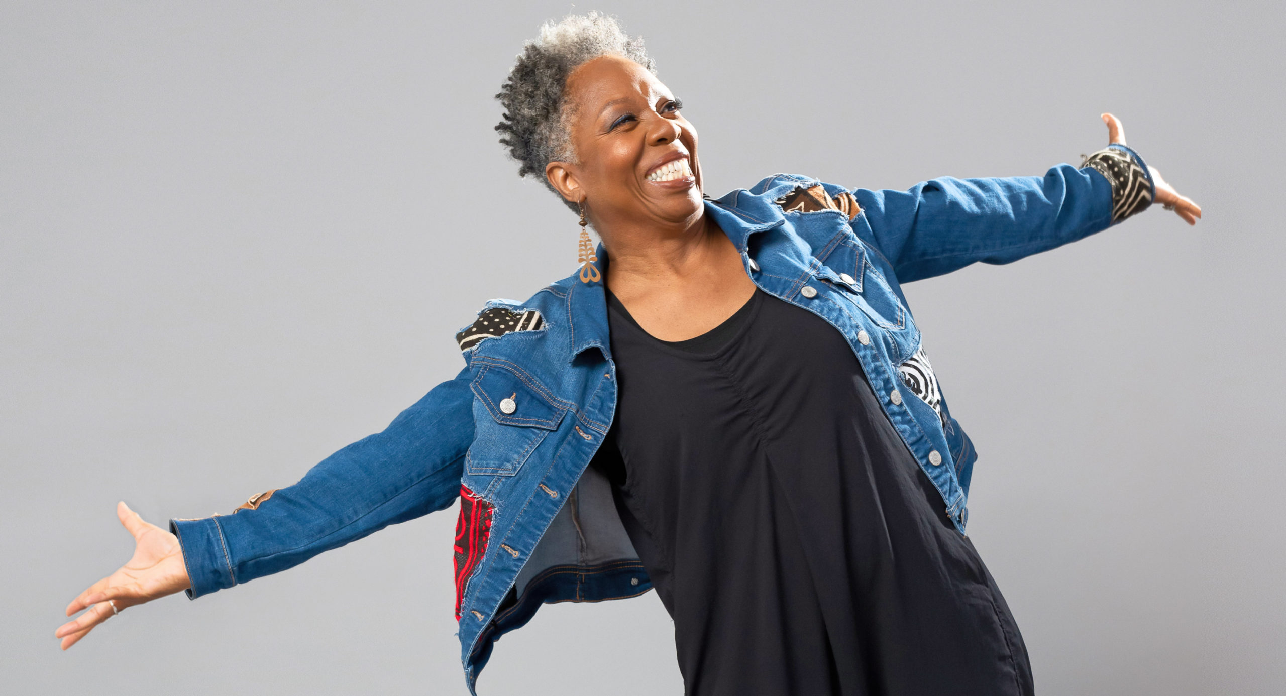 Against a grey backdrop, Jawole Willa Jo Zollar smiles widely, arms thrown open to either side. She wears a denim jacket with colorful patches over a simple black dress and a pair of striking light brown geometric earrings.