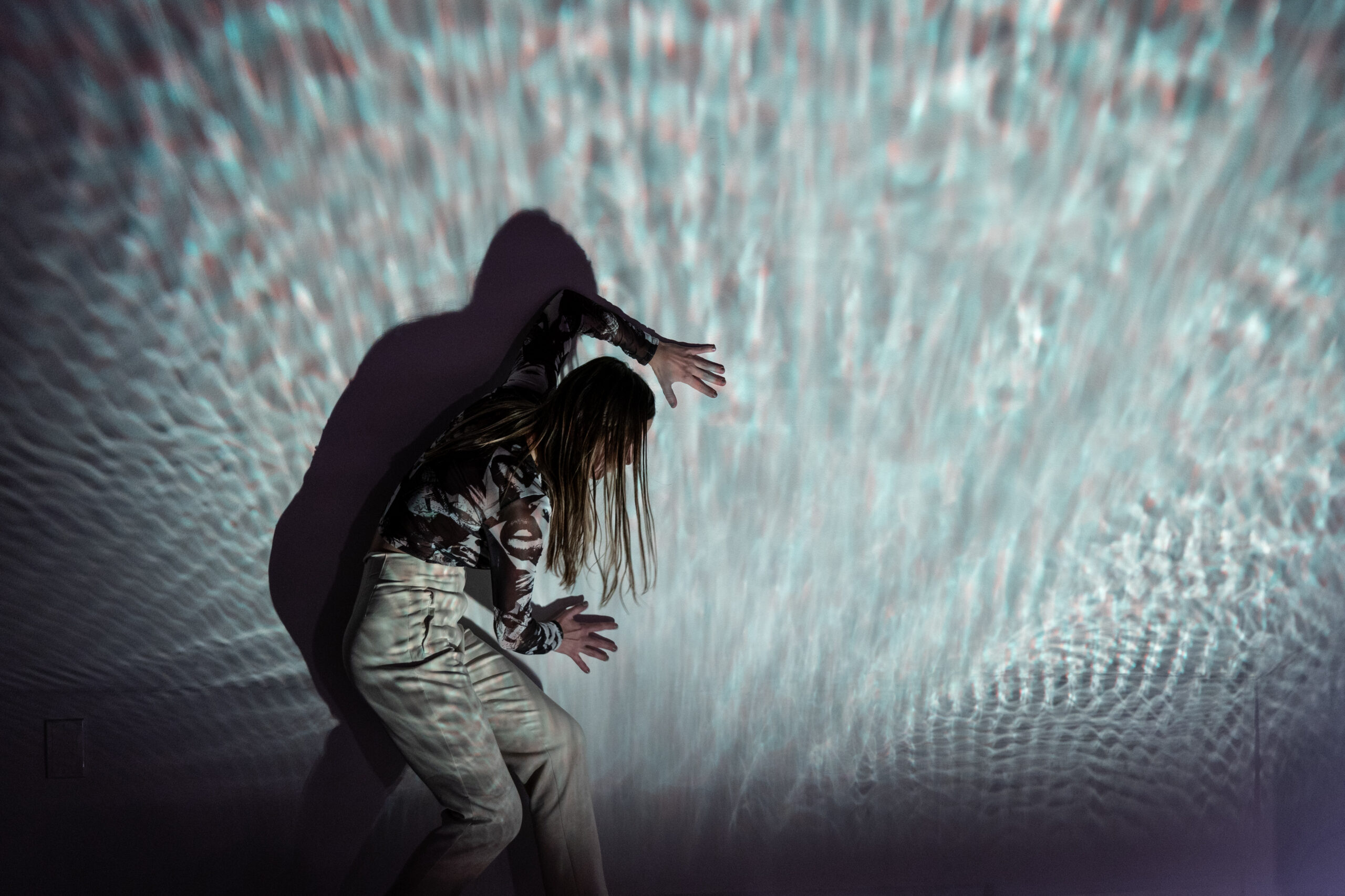 A dancer stands in profile against a wall, slightly stooped over as they press their palms against it, head turned toward it. A projection refracts from and over the dancer, who wears light colored clothing.