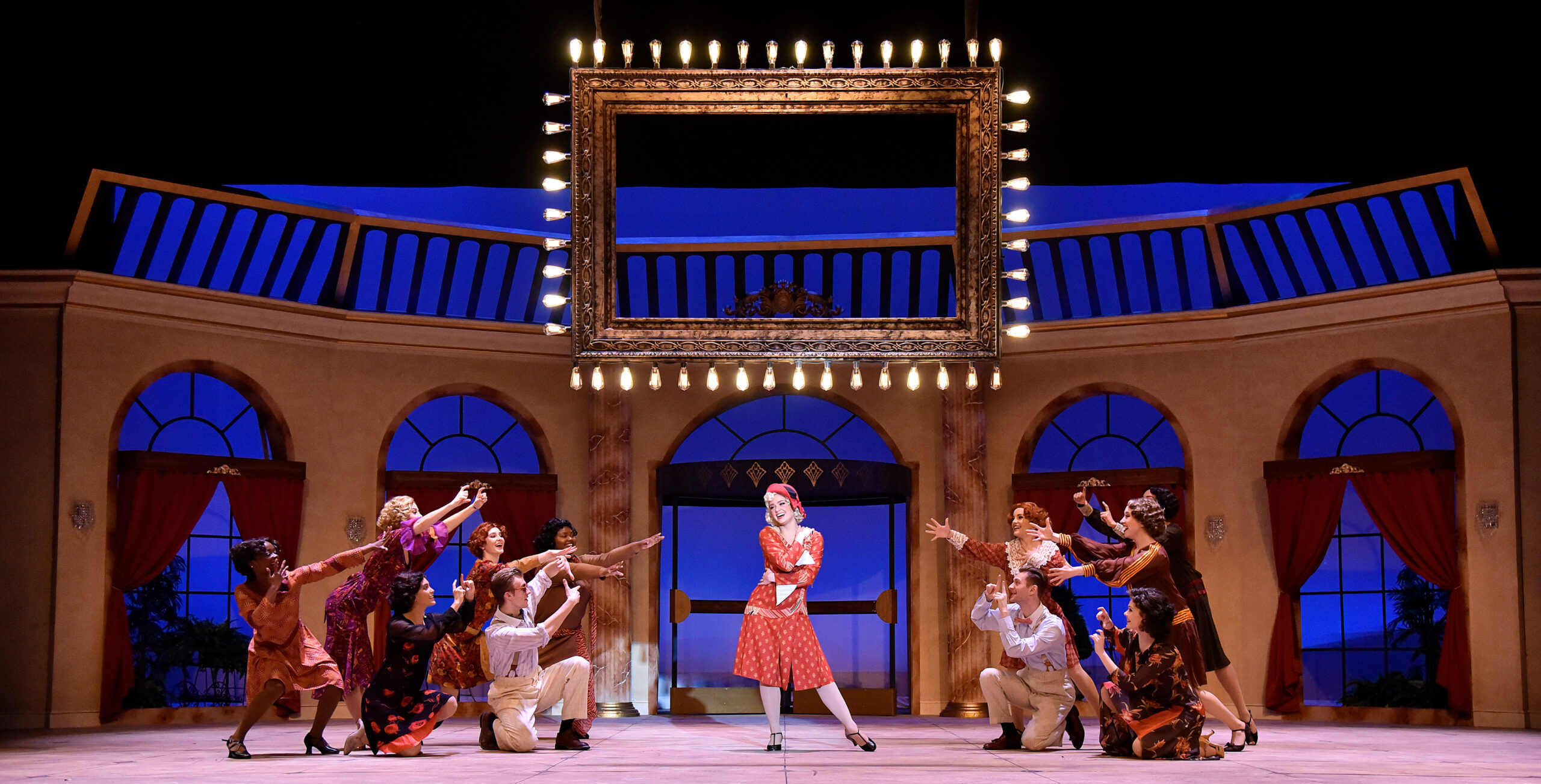 a large group of performers on stage, on female standing in the middle and the others around her and acting as if they're taking her photo