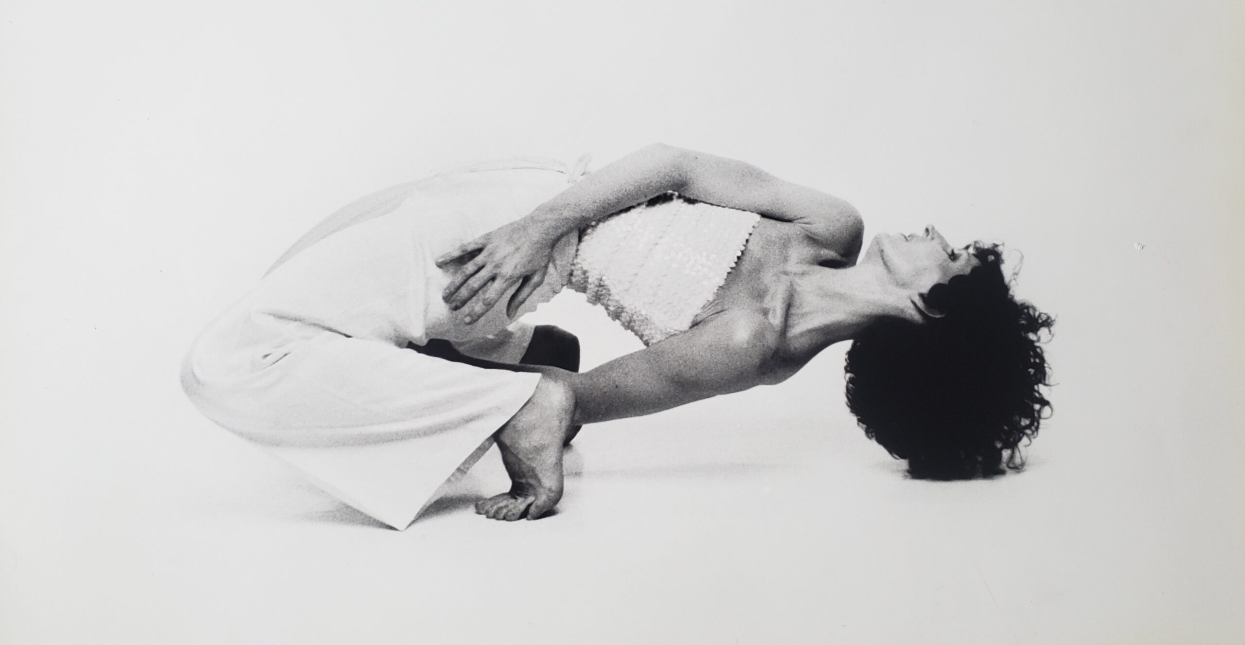 A black and white archival image of Jennifer Muller. She is in profile, serenely hinging back so her loose hair brush the floor, her body arcing almost parallel to the ground. Her upstage arm drapes in a longer curve over her torso so the hand rests over her downstage hip.
