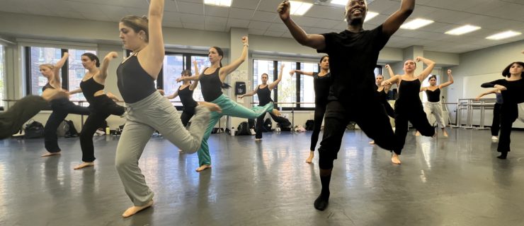 A group of students hop onto their right legs, their left arm overhead. They are led by a teacher demonstrating the same movement.