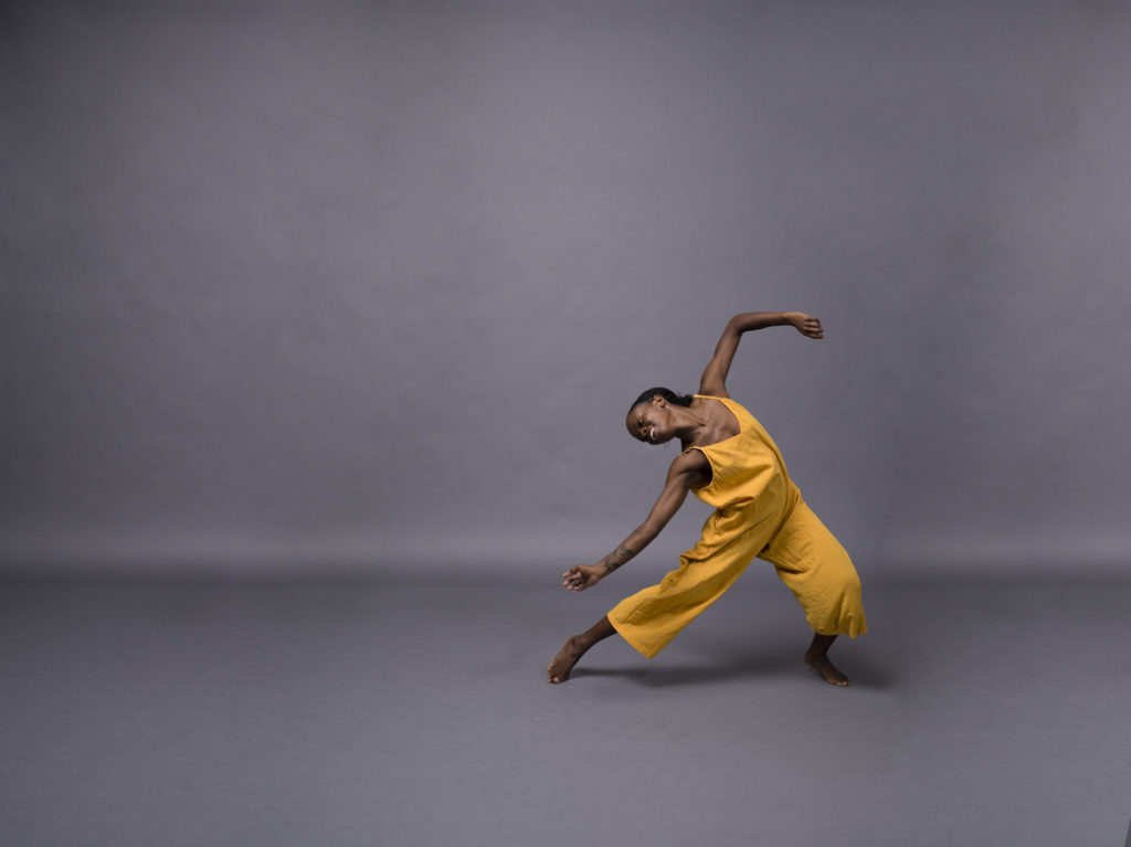 Against a grey backdrop, Simone Stevens, wearing a yellow gold jumpsuit, smiles joyfully as she moves through a deep plié, almost lunging. Her right arm curves to match her extended leg, while the left bends gently overhead. She arches right and back.