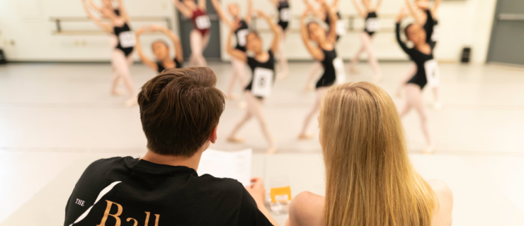 two adults sitting at a table watching a group of female dancers audition
