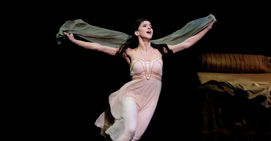 Mayara Magri is captured mid-run, arms outstretched to the sides as the cloak she holds in her hands billows behind her. Her costume is a pale peach dress over pink tights, reminiscent of a nightgown. Her hair is loose around her shoulders, a look of mingled despair, fear, and hope on her face.